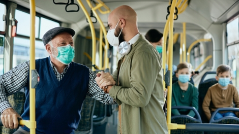 Ein jüngerer Mann mit Maske hilft einem älteren Mann mit Maske im Bus. Quelle: AdobeStock/drazen